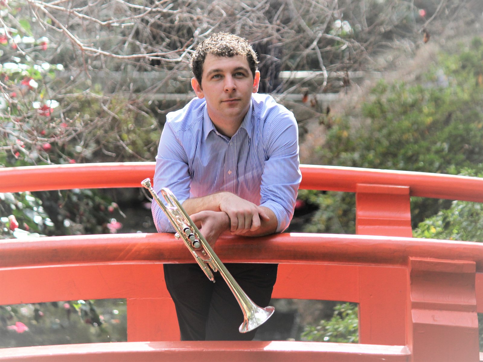 Portrait of Dylan Prothro holding his trumpet, leaning on a red garden bridge